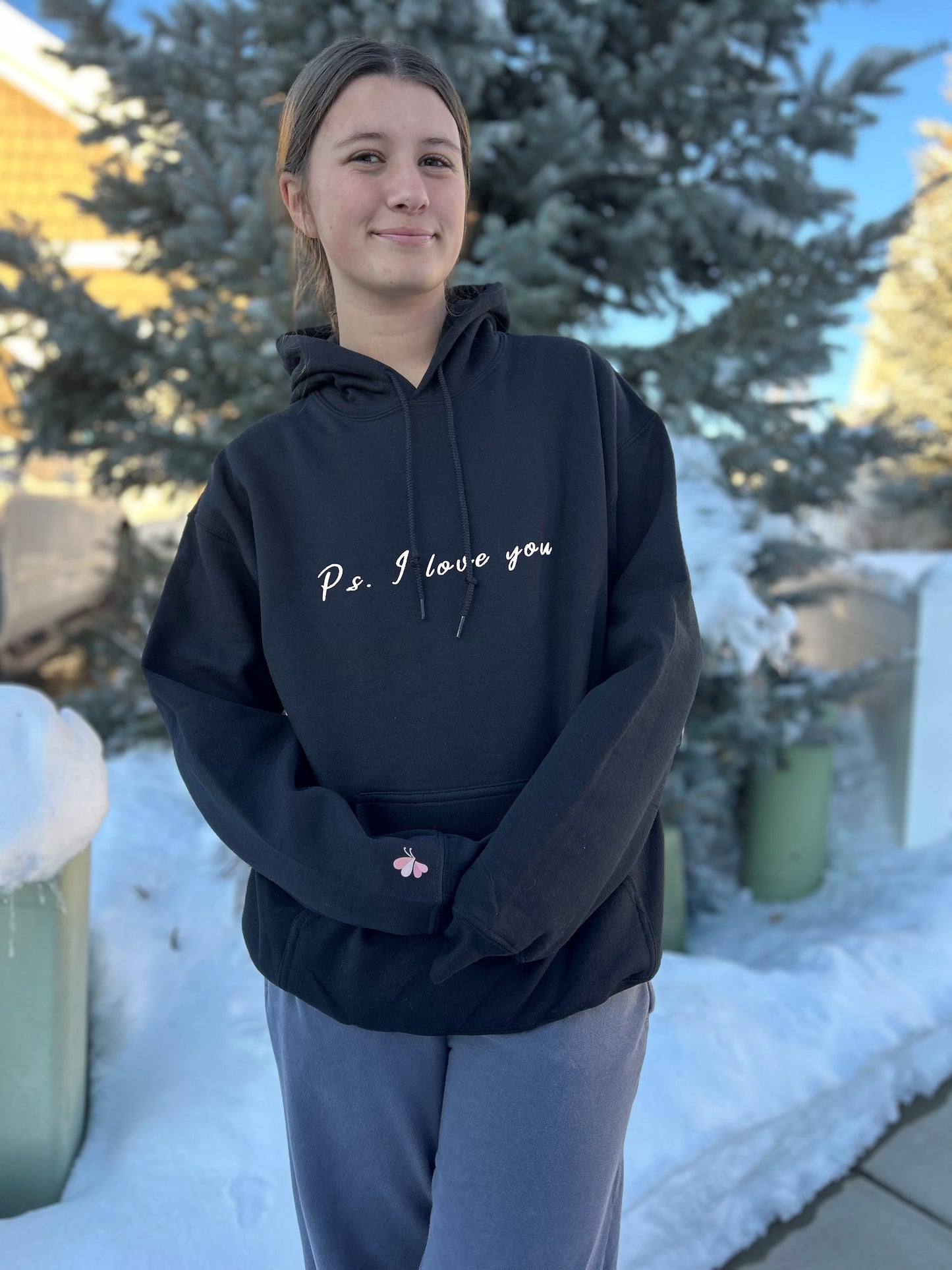 PS I Love You Mental Health Hoodie - a girl wearing a black hoodie with words that read PS I Love You.  There is snow and a pine tree in the background.