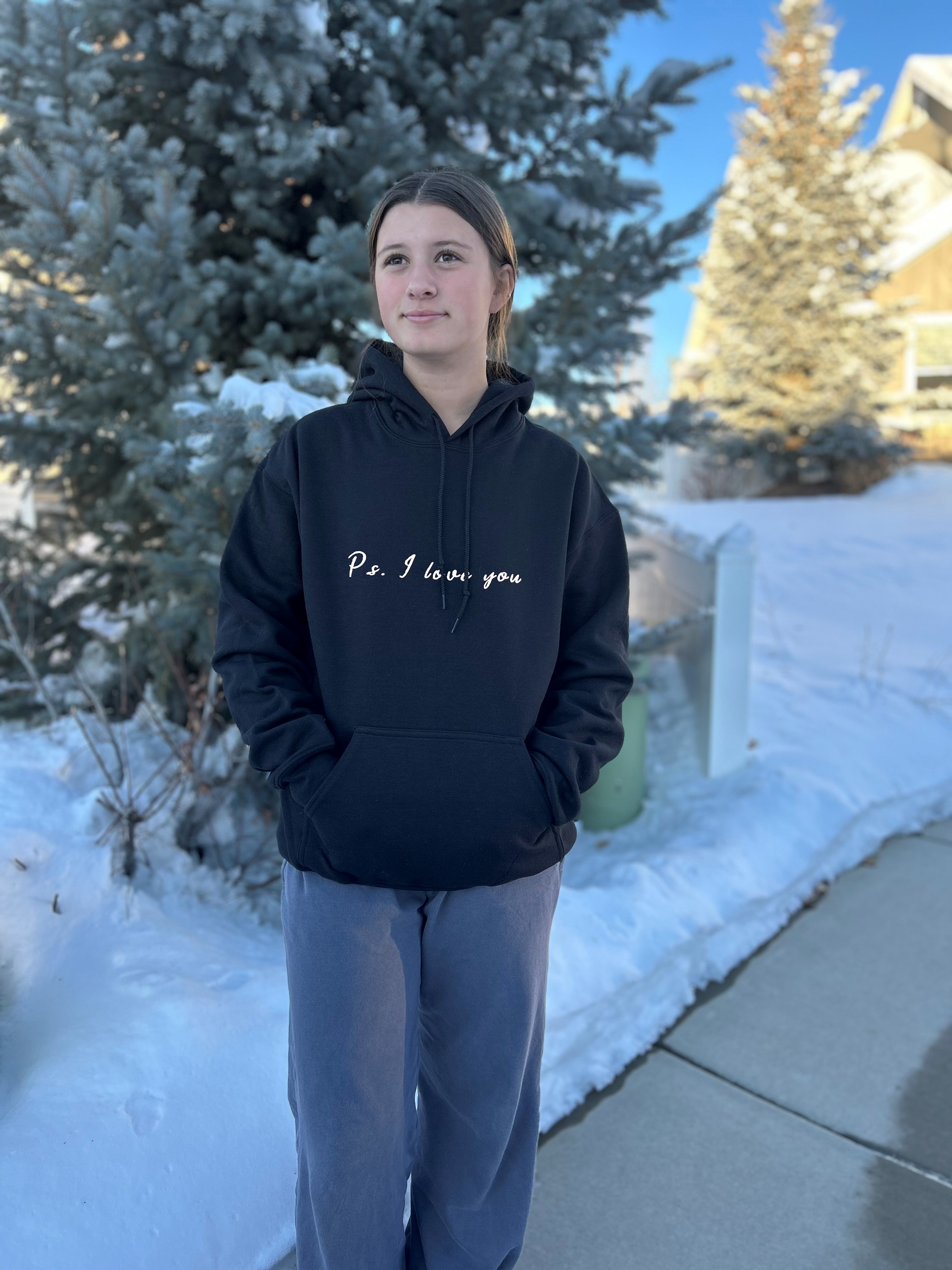 PS I Love You Mental Health Hoodie - a girl wearing a black hoodie with words that read PS I Love You.  There is snow and a pine tree in the background.